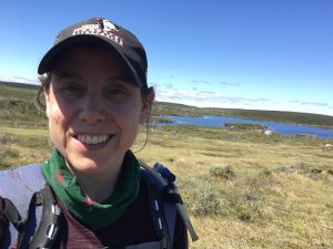 Amy-Lee looks at the camera and smiles, she is wearing a baseball cap to shield her eyes from the sun. A flat, grey-green landscape stretches behind her toward a body of blue water