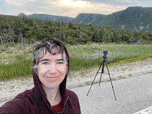 Robin smiles into the camera with a black hoodie on. In the background is a black tripod set up on sand, with evergreen trees and mountains further in the background